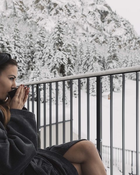 Bilder aus der Sportalm Frau im Bademantel trinkt Heißgetränk auf Balkon mit verschneitem Bergblick