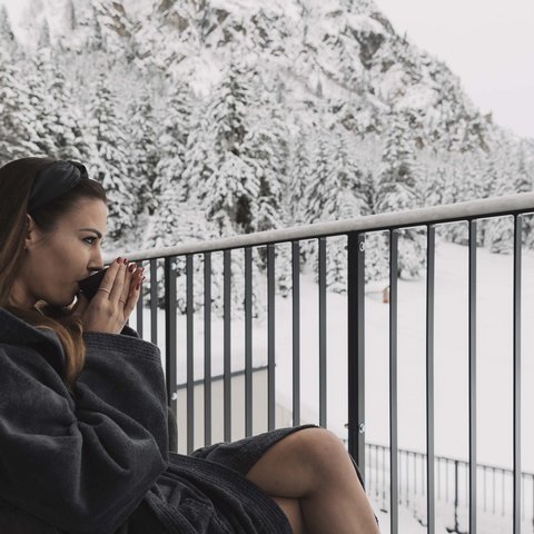 Bilder aus der Sportalm Frau im Bademantel trinkt Heißgetränk auf Balkon mit verschneitem Bergblick