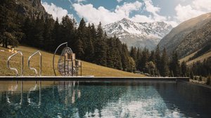 Hiking hotel in Pitztal: nature adventurers, ahoy! Mountain landscape with pool and stools in front of pine forest and snow-covered mountain